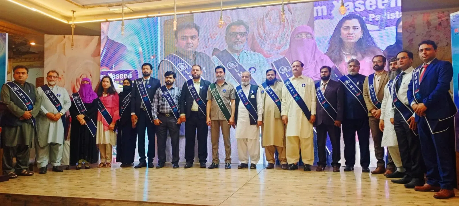 A long row of members wearing LMHI sashes on a stage before a large screen, with Dr. Mir Zahed fourth from the right.