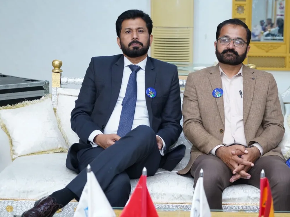 Dr. Mir Zahed seated on an ornate white and gold sofa beside another participant, behind a table of small flags.