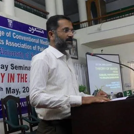 Dr. Mir Zahed lecturing at a podium. The slide behind him reads “Theory of Miasm, from Samuel Hahnemann to Rajan Sankaran”.